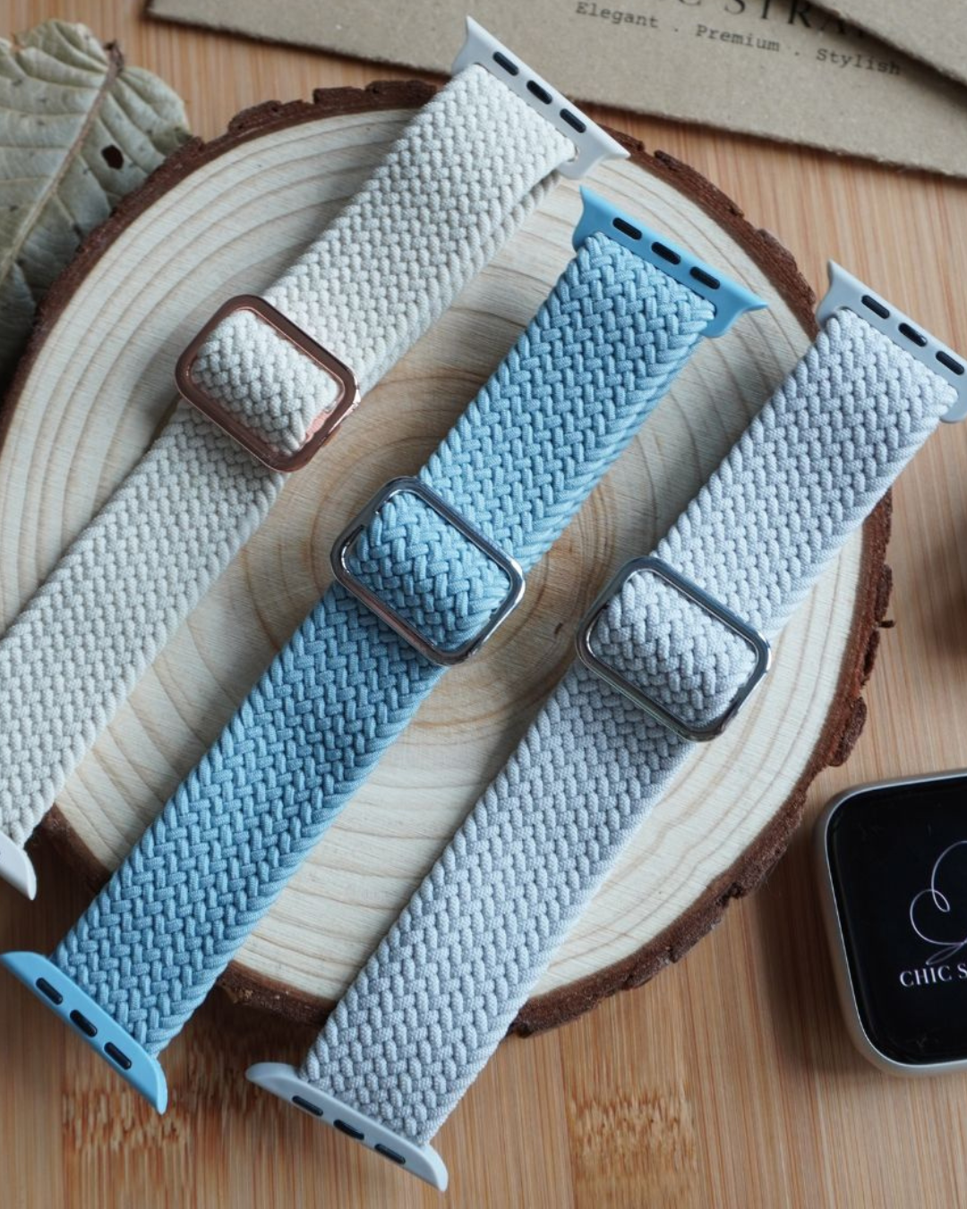 Three braided watch straps in different colors on a wooden surface with a phone displaying 'Chic Straps' branding.